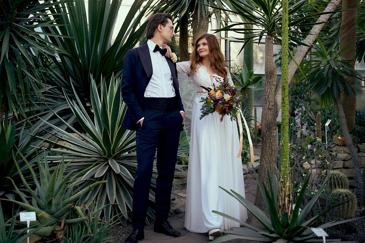 Panna młoda w białej sukni trzyma kolorowy bukiet i uśmiecha się do pana młodego w smokingu, gdy stoją razem w otoczeniu bujnych, zielonych roślin tropikalnych w szklarni.