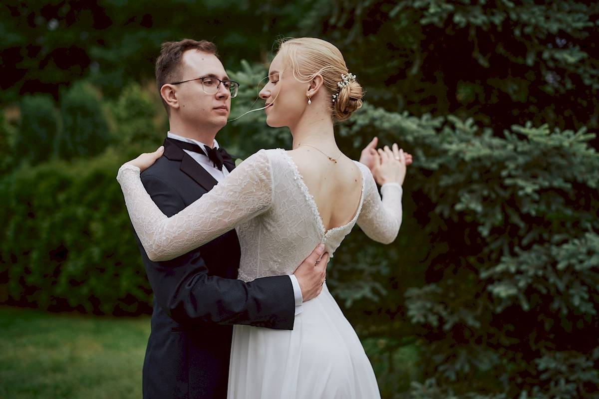 Panna młoda i pan młody w formalnych strojach tańczą na świeżym powietrzu w pobliżu zieleni, a panna młoda żartobliwie trzyma w ustach kawałek trawy i oboje patrzą na siebie.