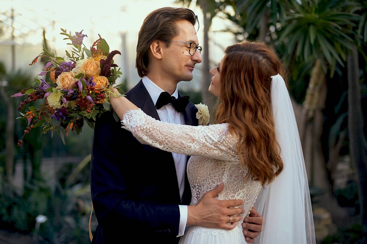 Panna młoda i pan młody w formalnych strojach ślubnych obejmują się i patrzą sobie w oczy na świeżym powietrzu; panna młoda trzyma kolorowy bukiet i ma na sobie koronkową suknię z welonem. Otacza ich bujna zieleń.